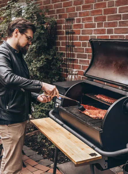 A man in a leather jacket turning spare ribs on a MOESTA Sheriff pellet grill with bamboo side shelf