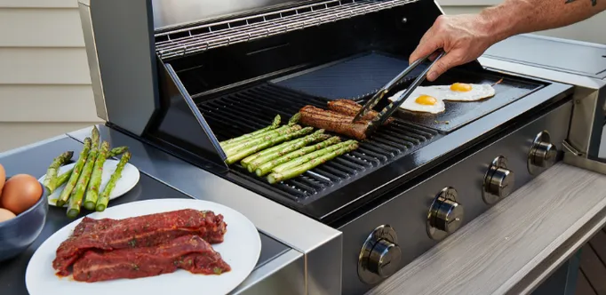 Grill grate with meat and asparagus being flipped with tongs, with raw meat and eggs nearby
