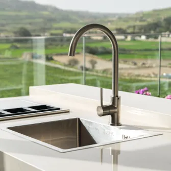 Sink and faucet of an outdoor kitchen on a balcony with glass railing and idyllic green landscape behind