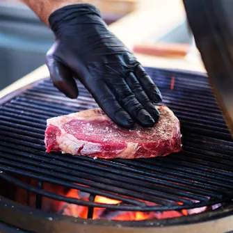 Hand mit Handschuhe die ein rohes Steak auf einen befeuerten Grillrost legt