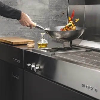 Hand schwenkt eine Pfanne mit Grillgemüse über der Herdplatte einer Flammkraft Outdoorküche