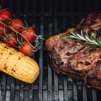 Close-up of grilled tomatoes, a corn cob, and a steak on a grill rack