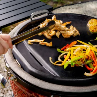 Close-up of the grill surface of a Kamado Joe grill with meat and bell pepper strips being turned with tongs