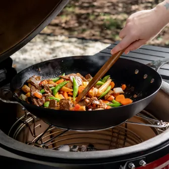Cast iron pan in a Kamado Joe, stirring vegetables and meat with a wooden spoon